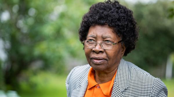 Nellie Suffolk stands in her garden looking away from the camera. She is wearing glasses and a bright orange shirt with a dogtooth print blazer.