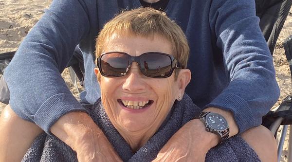 Glenys Smith sits on the beach, her husband Ralph, is embracing her from behind while sitting in a camping chair.