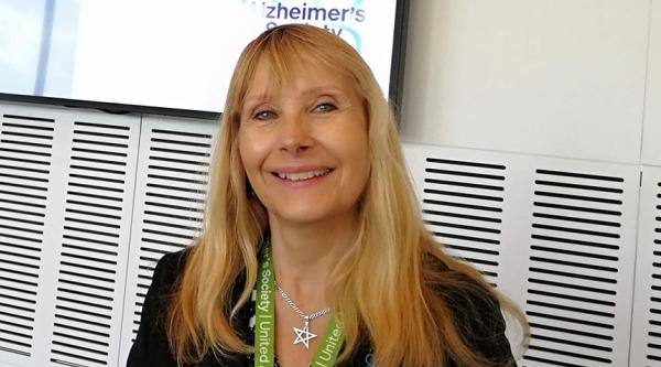 Lorraine Brown smiling and stood in front of a TV screen wearing a green lanyard