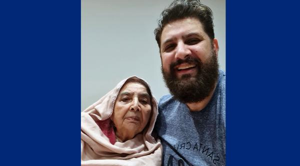 Matt is smiling and wearing a blue t-shirt. He has a black hair and a beard. His mum Kaniz is next to him and wearing a pale pink headscarf.