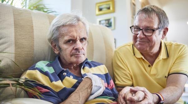 A man in a yellow top holds the hand of a man sat in a beige sofa wearing a colourful stripy top