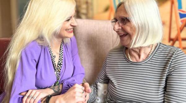 Kerri is talking to a care home resident and holding their hand. She's wearing a purple blouse and has long blonde hair