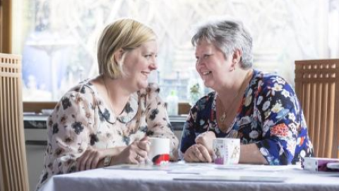 Two women sit closely together at a table with cups of tea.